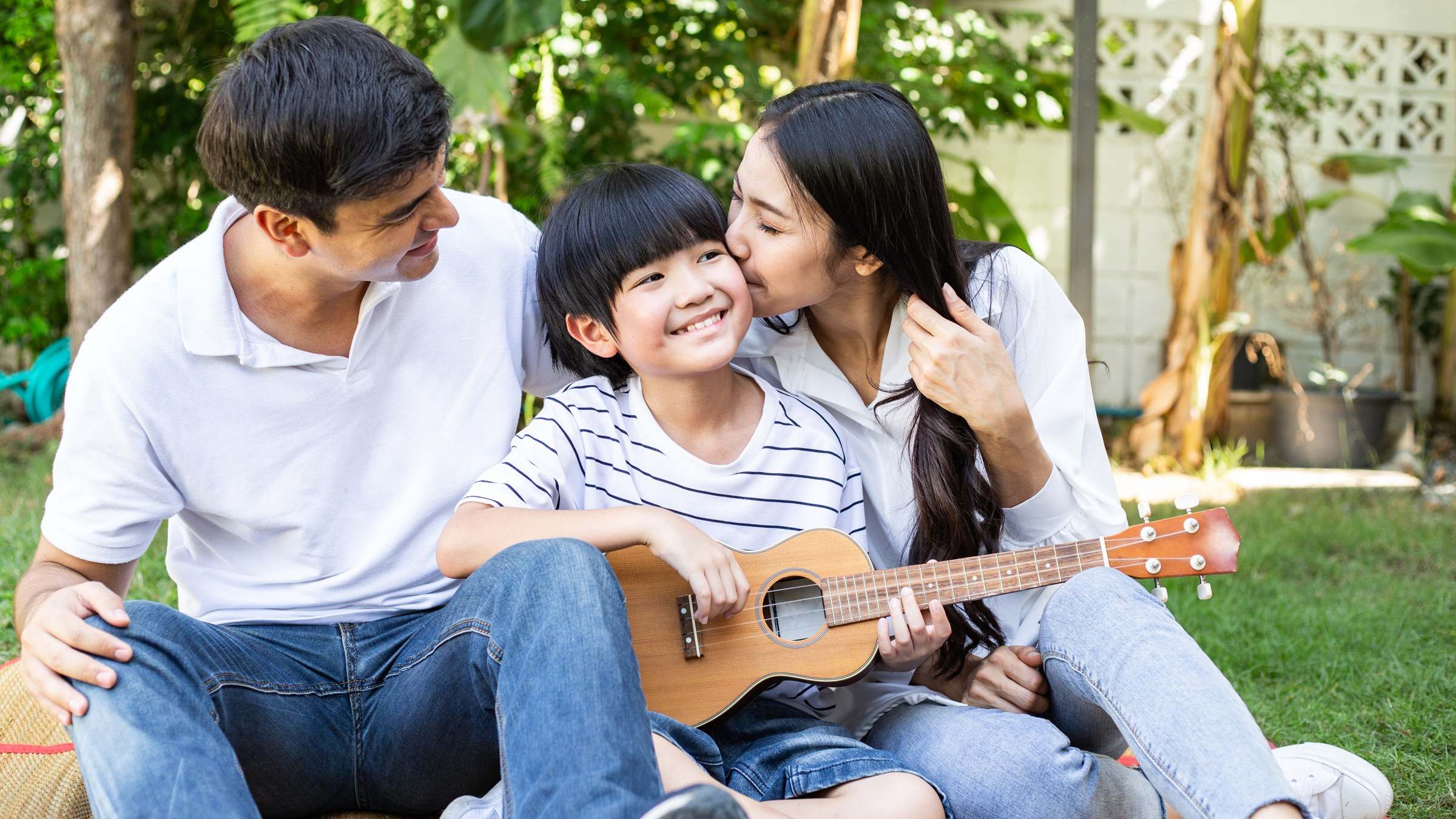 Asian family relaxing together in their garden, the son playing a ukulele