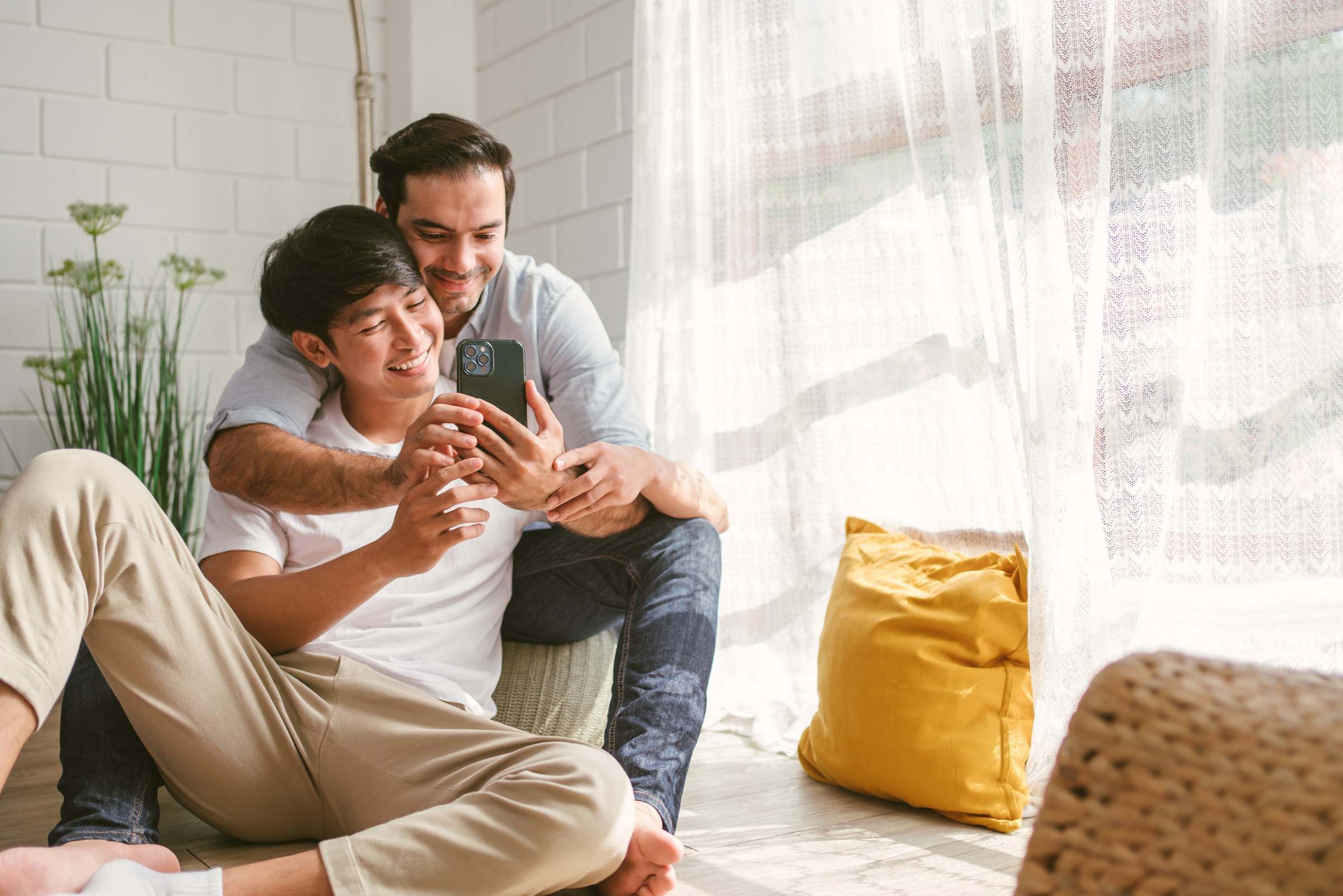 Asian and White male couple smiling at a phone together by a sunny window
