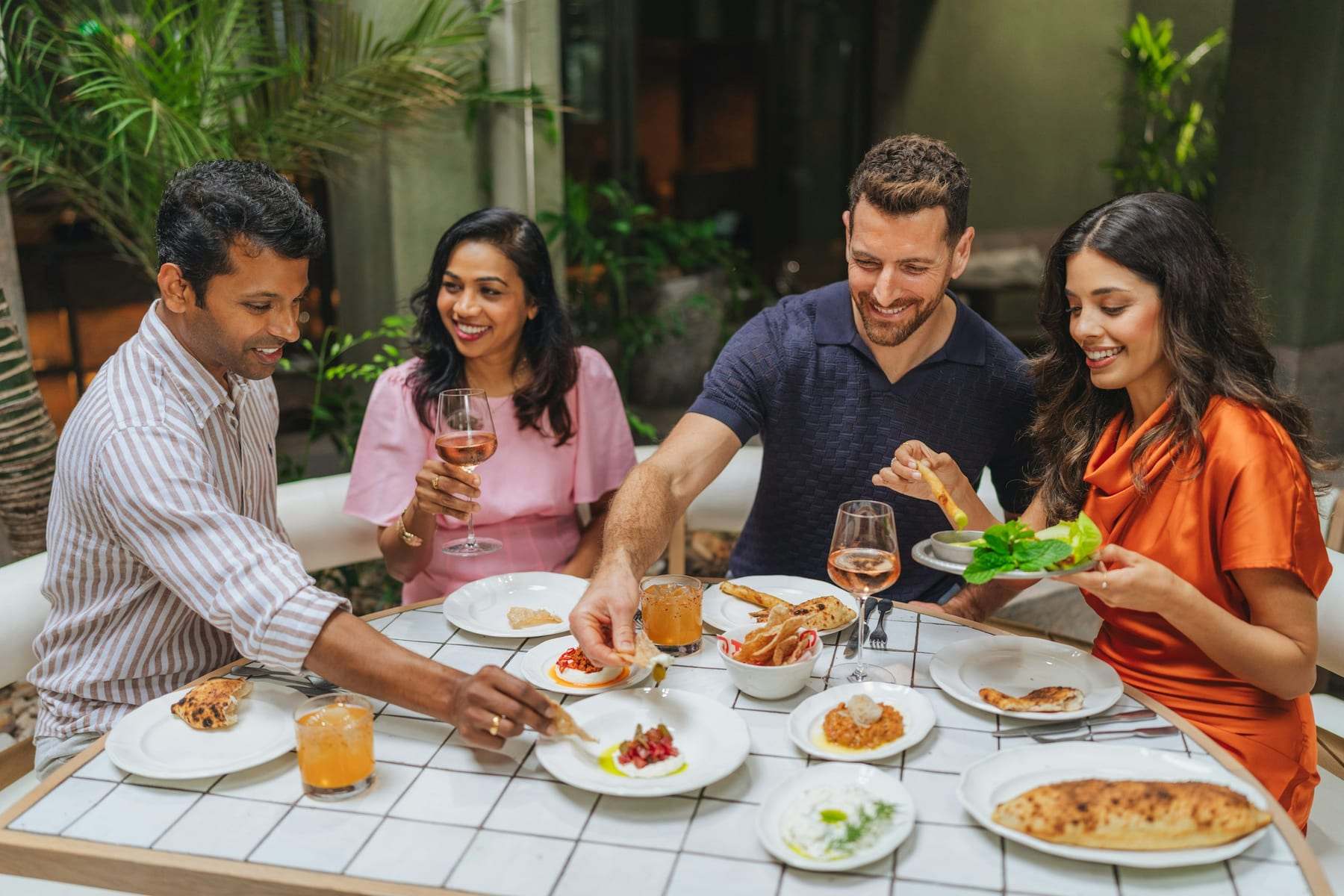 Friends sharing small plates and wine at a tiled table on a leafy terrace
