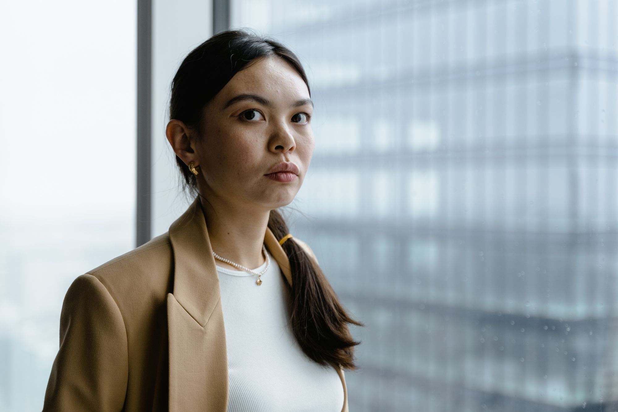 Asian businesswoman in a camel blazer standing by a tall office window
